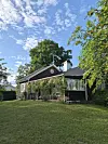 Friliggande villa med veranda, stor gräsmatta och träd i soligt väder.