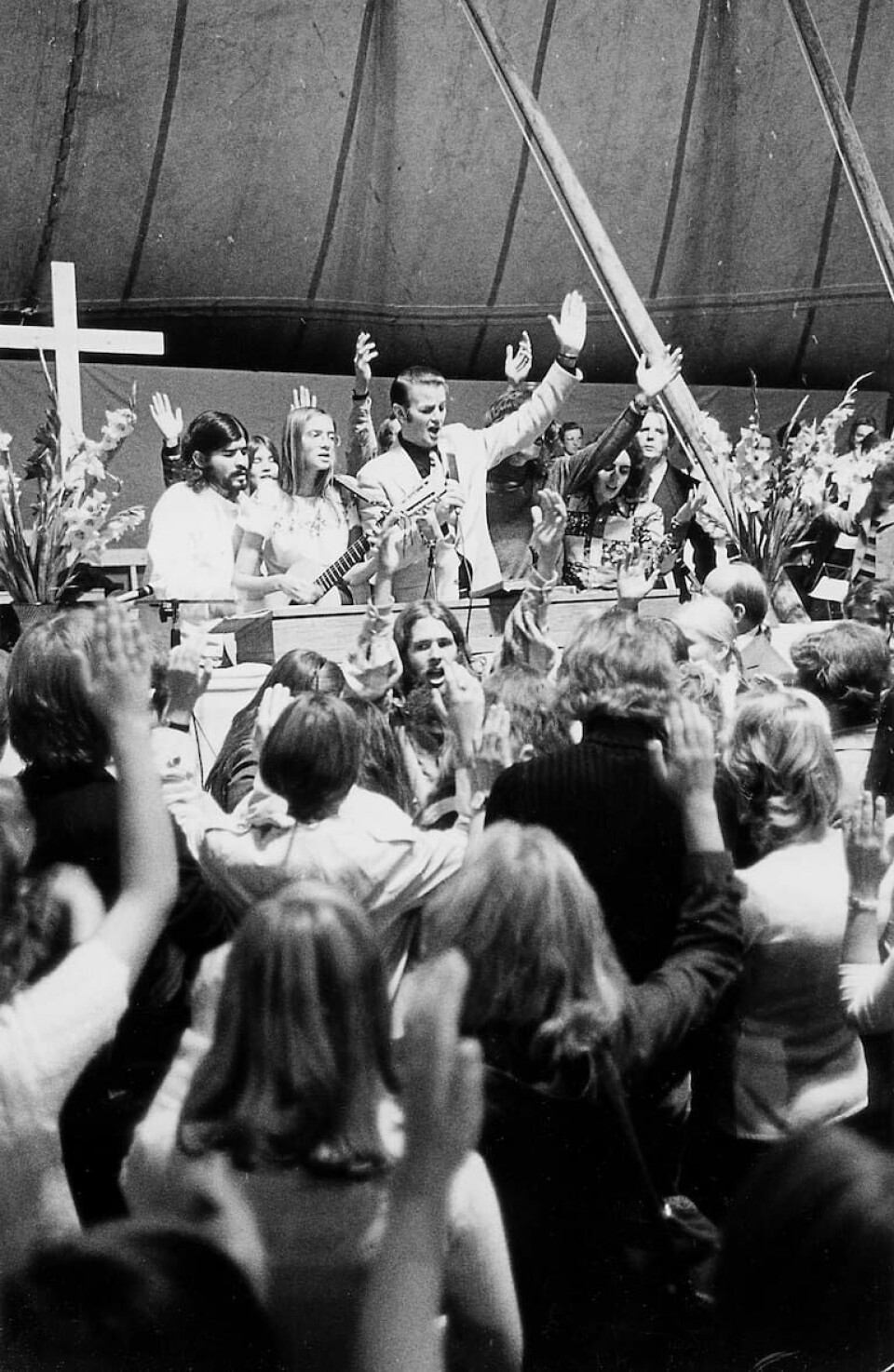 Stanley Sjöberg i talarstolen och hippievangelisten Lonnie Frisbee nedanför bland mötesbesökarna, i mitten av bilden. Under pingströrelsens Nyhemsvecka vid besöket i Sverige 1971. Jesusfolkets besök på Nyhemsveckan 1971
